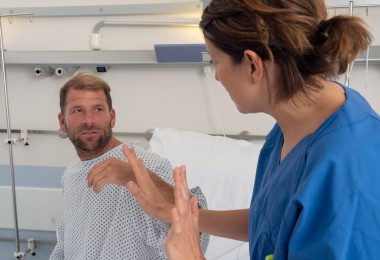 Ein Mann in Krankenhauskleidung sitzt aufrecht im Bett und schaut eine Pflegekraft an, die ihm gegenübersitzt und mit erhobenen Händen eine abwehrende Geste macht. Die Situation wirkt angespannt.