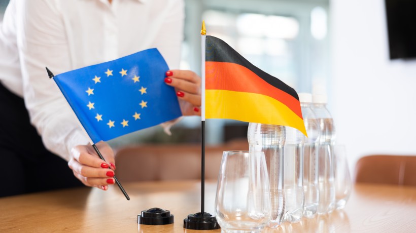 Eine Person im weißen Hemd mit rot lackierten Fingernägeln stellt zwei kleine Tischflaggen auf einem Konferenztisch auf: eine blaue Flagge der Europäischen Union mit gelben Sternen und eine schwarz-rot-goldene Flagge Deutschlands. Daneben stehen mehrere Wasserflaschen und leere Gläser. Eine Person im weißen Hemd mit rot lackierten Fingernägeln stellt zwei kleine Tischflaggen auf einem Konferenztisch auf: eine blaue Flagge der Europäischen Union mit gelben Sternen und eine schwarz-rot-goldene Flagge Deutschlands. Daneben stehen mehrere Wasserflaschen und leere Gläser.