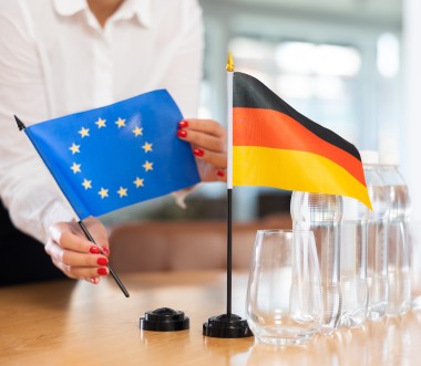 Eine Person im weißen Hemd mit rot lackierten Fingernägeln stellt zwei kleine Tischflaggen auf einem Konferenztisch auf: eine blaue Flagge der Europäischen Union mit gelben Sternen und eine schwarz-rot-goldene Flagge Deutschlands. Daneben stehen mehrere Wasserflaschen und leere Gläser. 