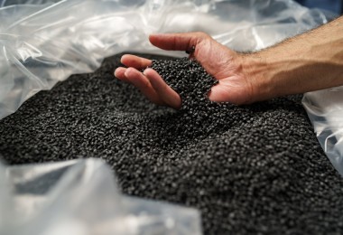 Eine Hand greift in einen großen Behälter mit schwarzen Kunststoffgranulaten und lässt einige der kleinen Körner durch die Finger gleiten.
